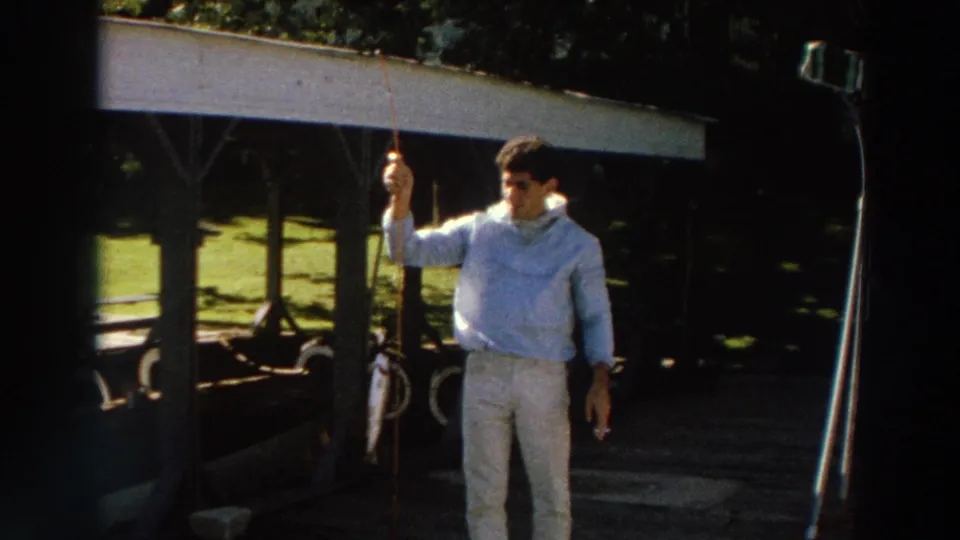In 1966 Madison home-movie footage, a boy in light clothing stands outdoors holding a fishing rod near a shaded roofed structure.