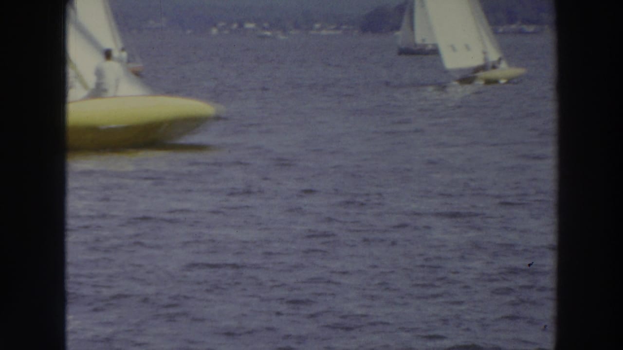 Yellow-hulled sailboat partly entering frame on a lake with other white-sailed boats in the distance.