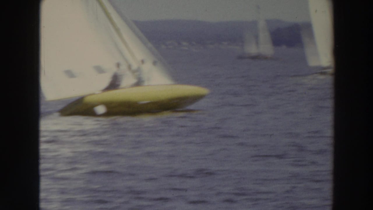 Yellow-hulled sailboat with two people aboard crosses the lake under a white sail.