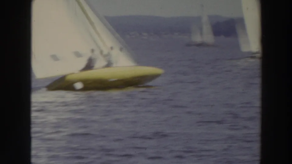 1966 Madison, Wisconsin 8mm frame of a yellow-hulled sailboat carrying two distant people, with other sailboats beyond it.