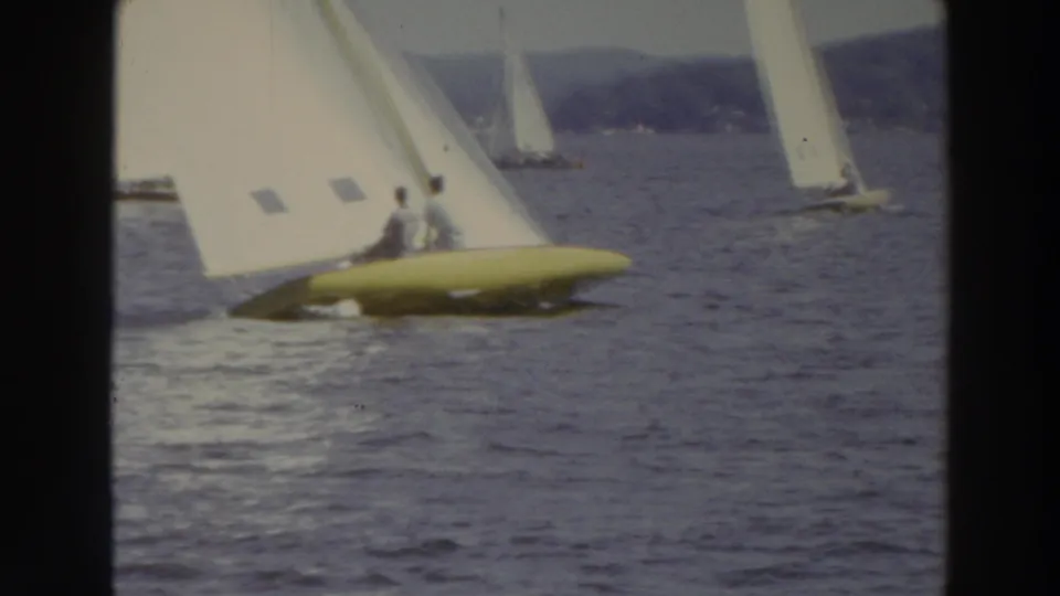 1966 Madison, Wisconsin 8mm frame showing a yellow-hulled sailboat in profile, two people aboard, and another sailboat near the shoreline.