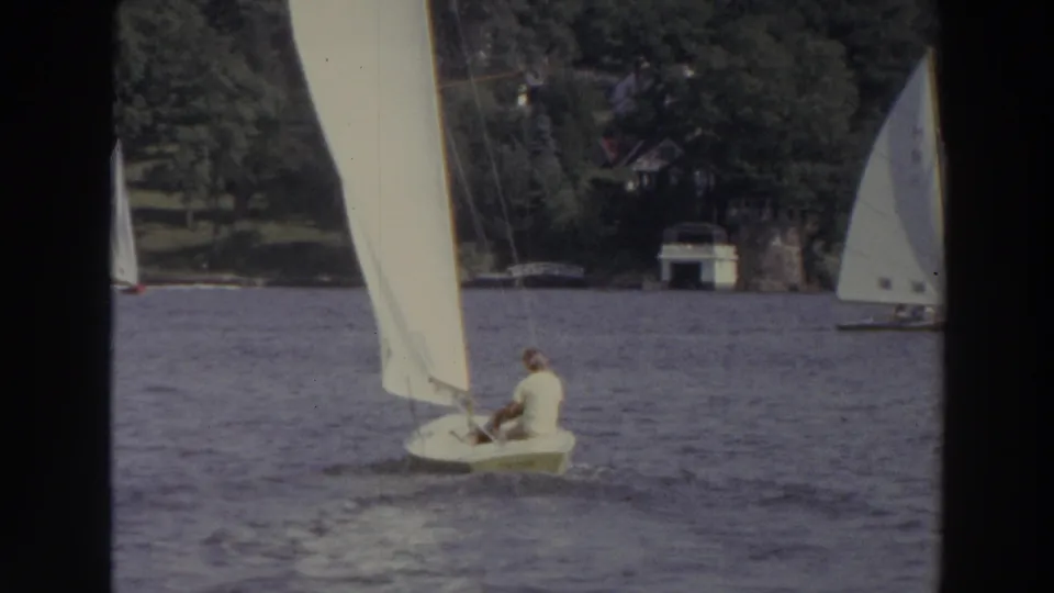 1966 Madison, Wisconsin 8mm frame with a seated adult sailor in a small dinghy, with other sails and wooded shore beyond.