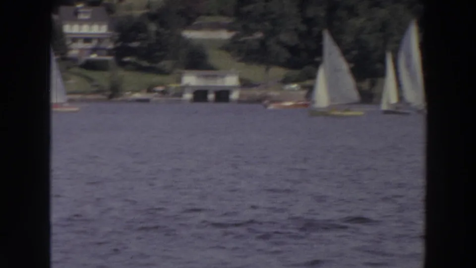 1966 Madison, Wisconsin home-movie frame showing several white sailboats moving across a lake near trees, lawns, docks, and houses.