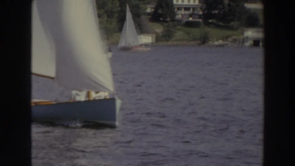 A nearby sailboat partly fills the frame as rippled lake water, distant white sails, trees, and lakeside houses remain visible beyond.