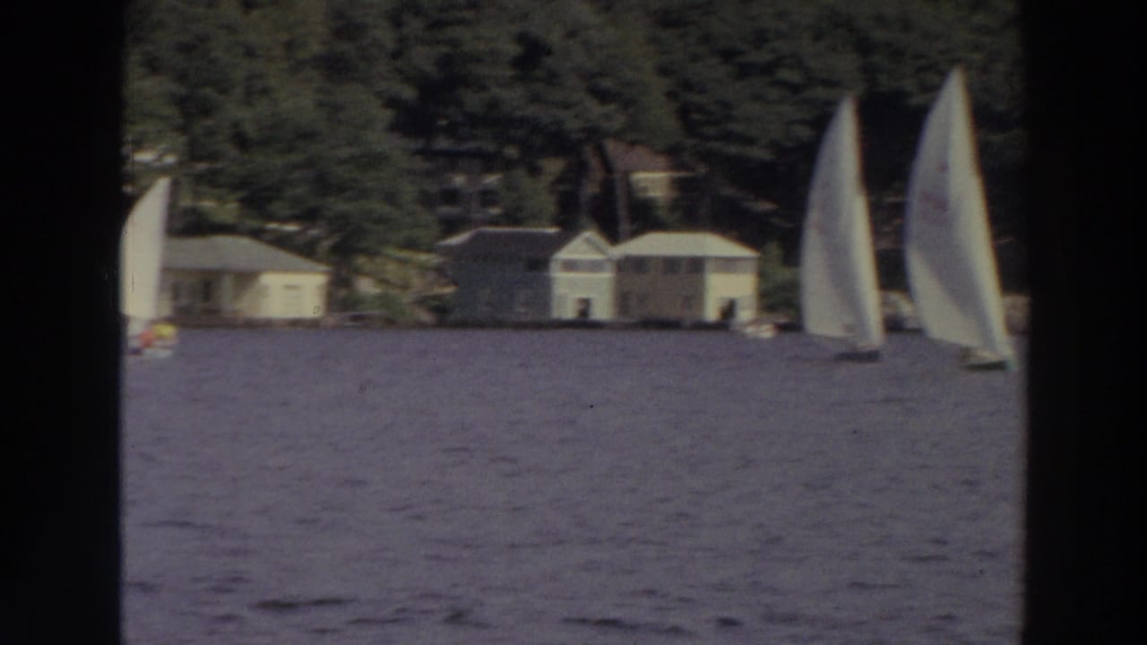 A close sailboat sail passes in front of distant lakeside houses.