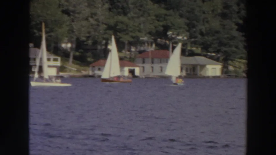 1966 Madison, Wisconsin 8mm frame showing sailboats on a lake with small shoreline houses and dense trees behind them.