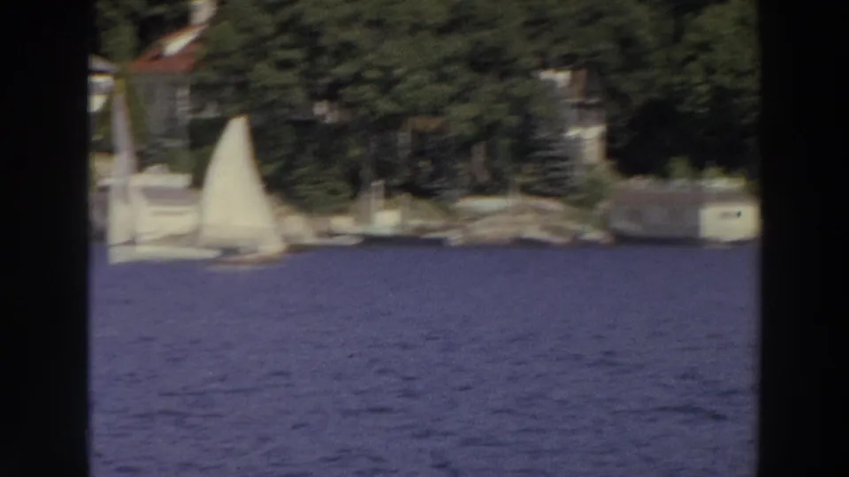 1966 Madison lake view with a white sailboat on blue water and houses partly visible among trees on the far shore.