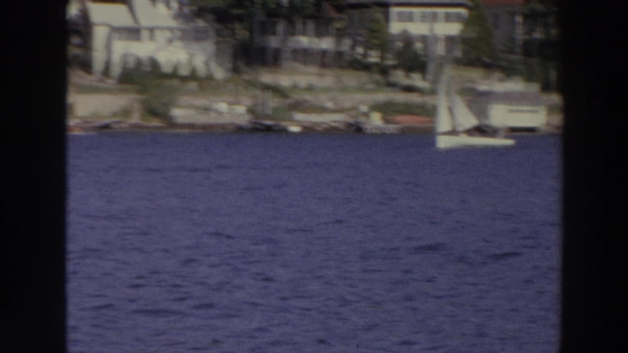 Distant white sailboats cross blue water in Madison, Wisconsin, with residential shoreline structures behind them.