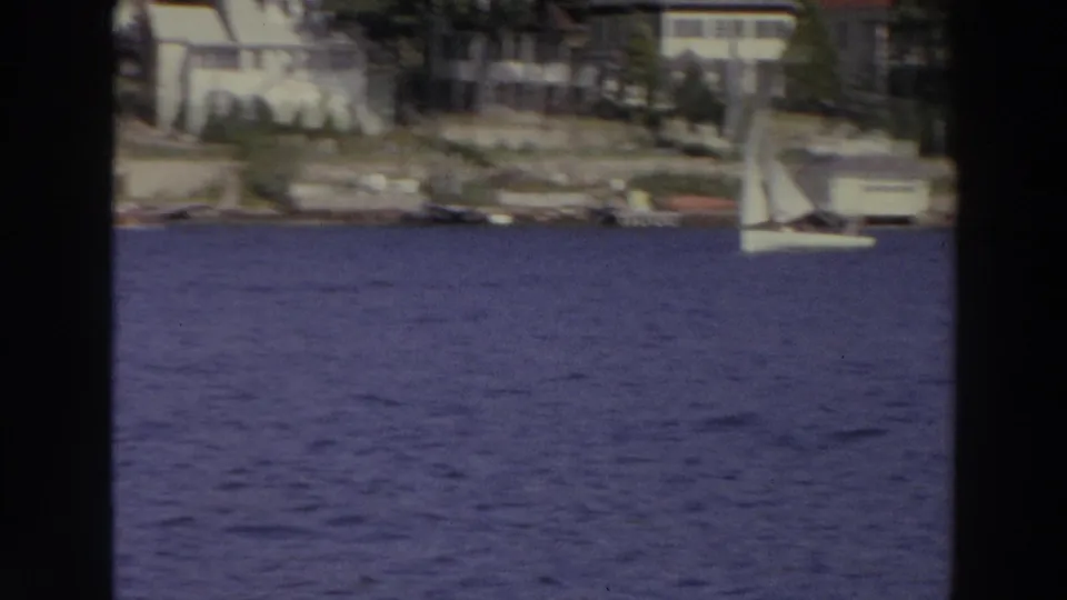 Distant white sailboats cross blue water in Madison, Wisconsin, with residential shoreline structures behind them.