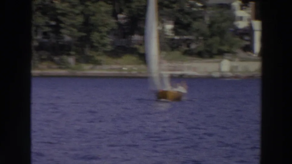 A soft, motion-blurred sailboat moves across a Madison lake, backed by trees and low shoreline structures.