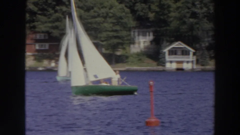 A white sailboat with incidental people aboard passes a red buoy on a Madison lake, with a boathouse and trees beyond.