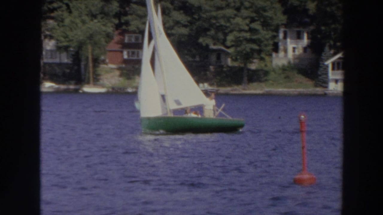 1966 Madison lake footage shows a green-hulled sailboat with white sails moving past a red buoy near a wooded shoreline.