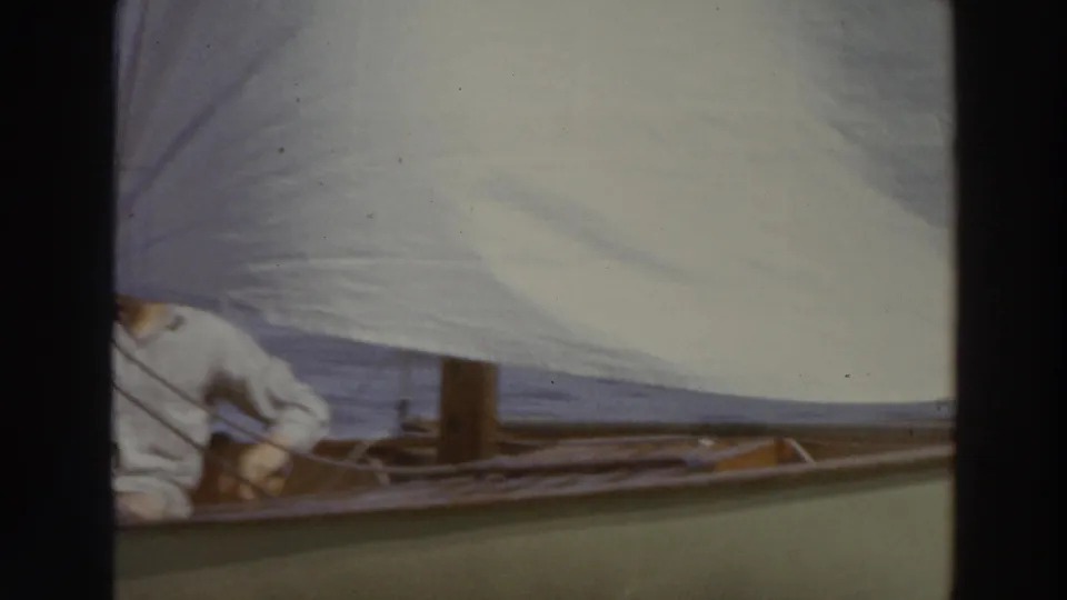 A crew member aboard a green-hulled sailboat sits beneath a white sail during 1966 Madison lake recreation footage.