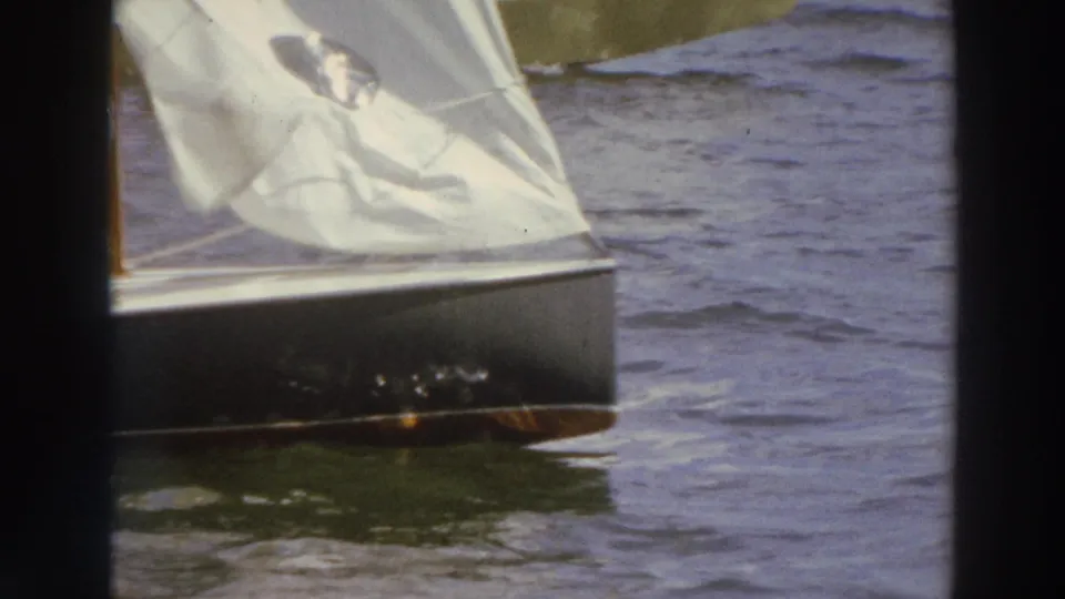 1966 Madison, Wisconsin 8mm frame showing the side of a small sailboat with a white sail on wind-ruffled lake water.