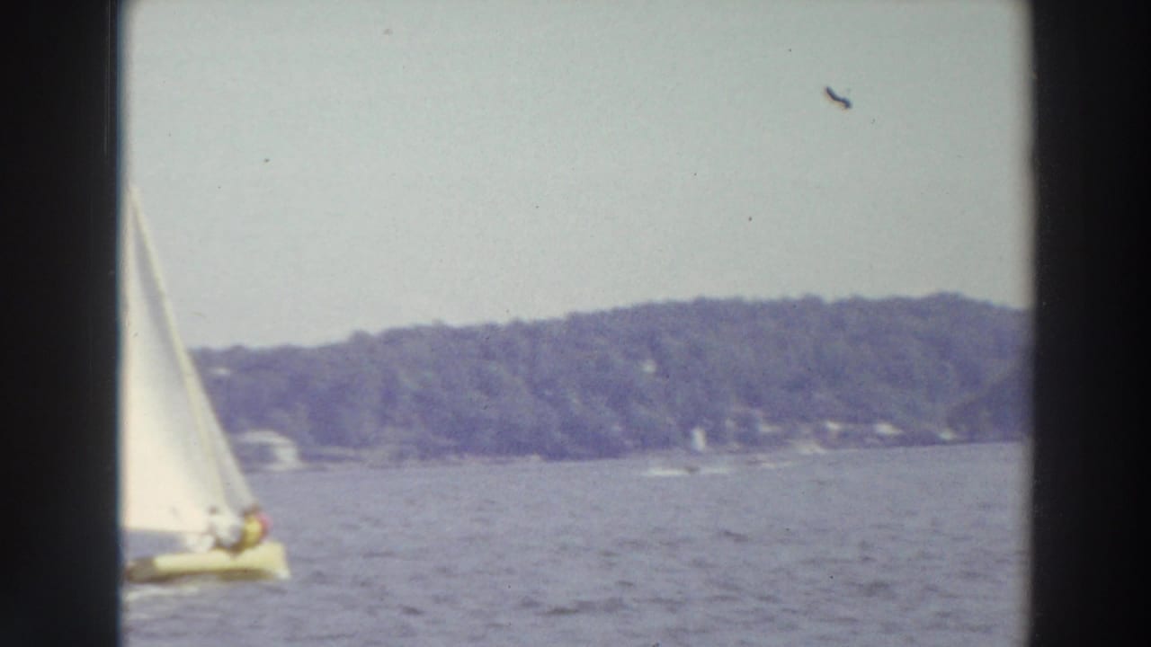 1966 Madison, Wisconsin sailing footage with a small white-sailed boat on open lake water and a tree-lined shore in the distance.