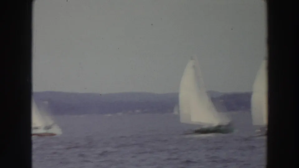 1966 Madison, Wisconsin home-movie frame with multiple white sails spread across the lake against a low wooded horizon.