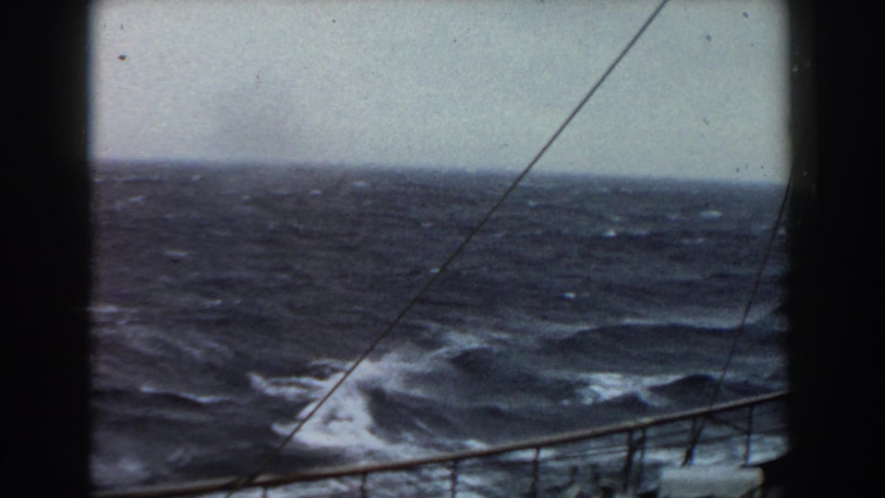 1959 8mm frame of dark rough seas and whitecaps viewed from a ship, with railing, rigging lines, overcast sky, and film gate edges visible.
