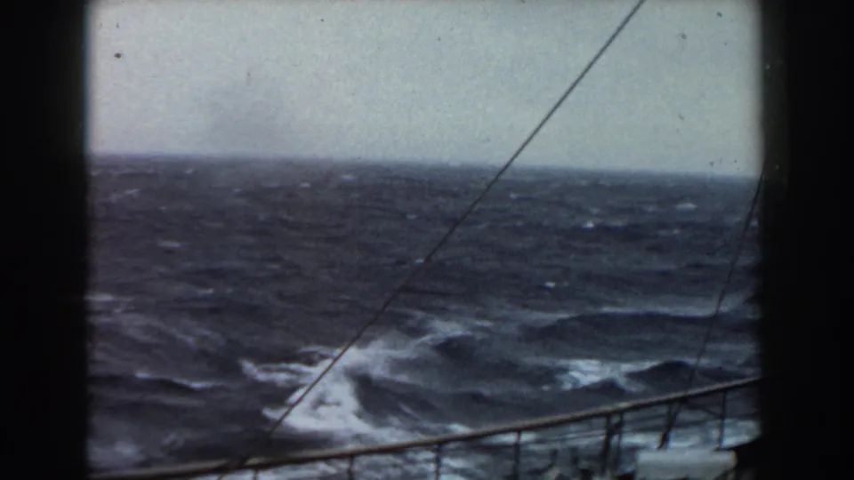 1959 8mm frame of dark rough seas and whitecaps viewed from a ship, with railing, rigging lines, overcast sky, and film gate edges visible.