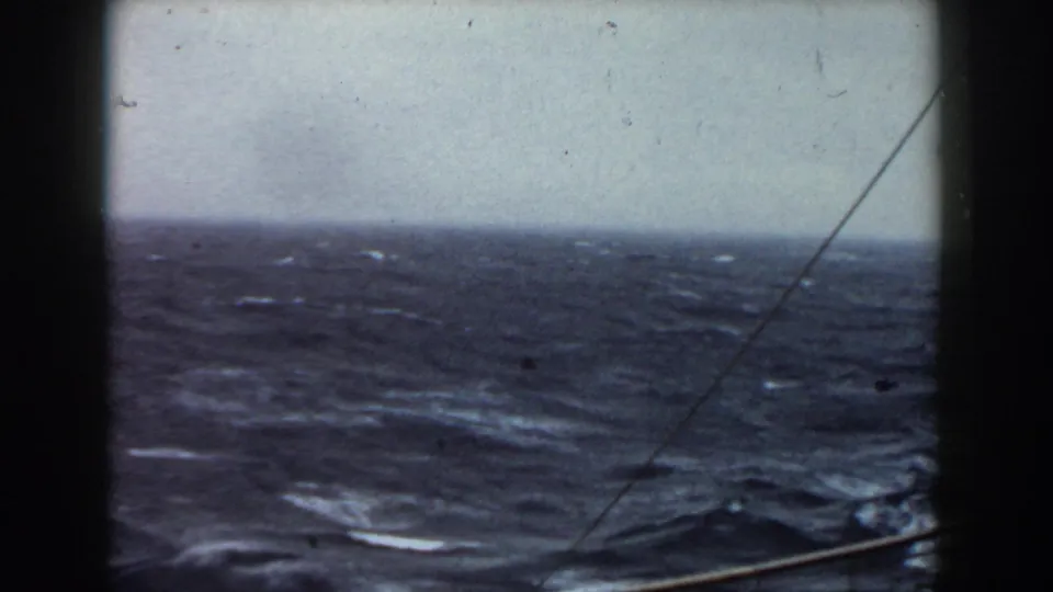 1959 8mm frame of choppy open water and scattered whitecaps seen from aboard a ship, with rigging lines and a low railing crossing the foreground.