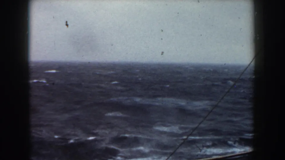 1959 8mm frame showing rolling dark water, whitecaps, an overcast horizon, and ship rigging crossing the view from a protected onboard vantage point.