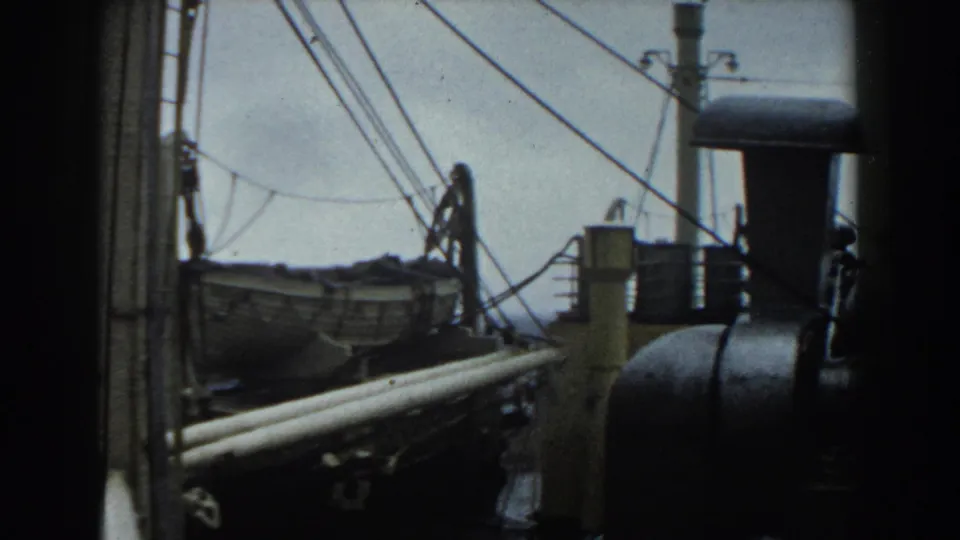 1959 shipboard view with a lifeboat, taut rigging, railings, and a dark rounded engine or vent structure on deck.