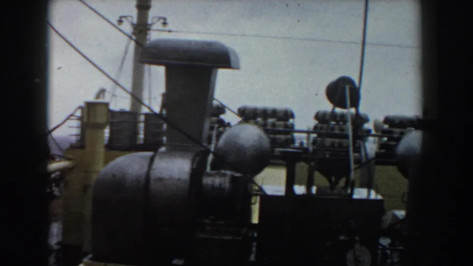 1959 8mm frame showing ship deck equipment, cables, vents, and ocean horizon from a traveler’s viewpoint.