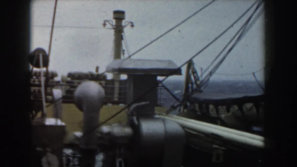 Handheld 8mm view across a ship deck with a small structure, rigging lines, rounded equipment, and open water.