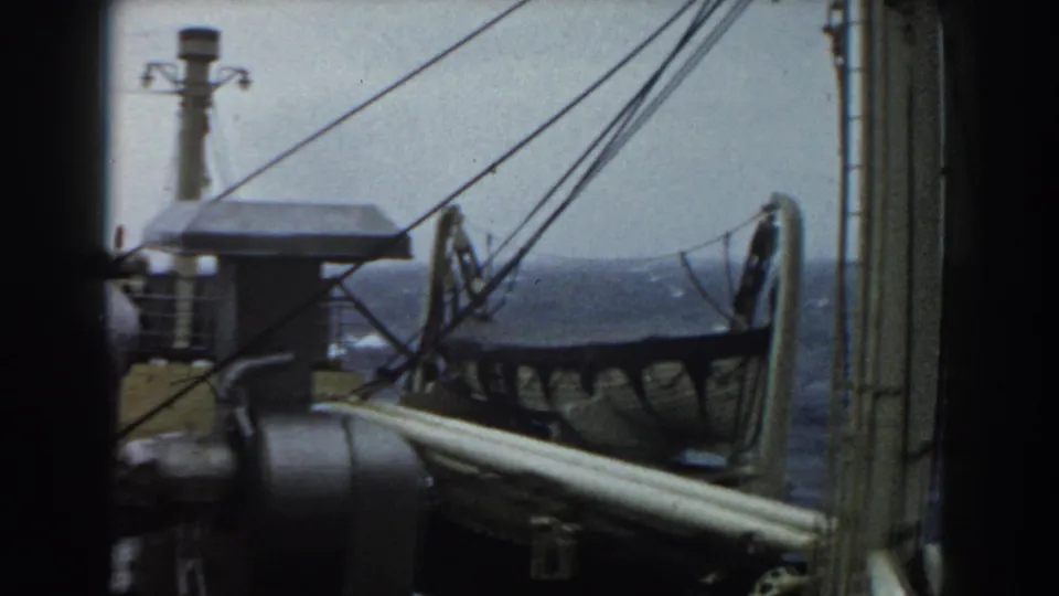 A shipboard frame showing railings, rigging cables, deck equipment, and a lifeboat-like form near the ocean view.