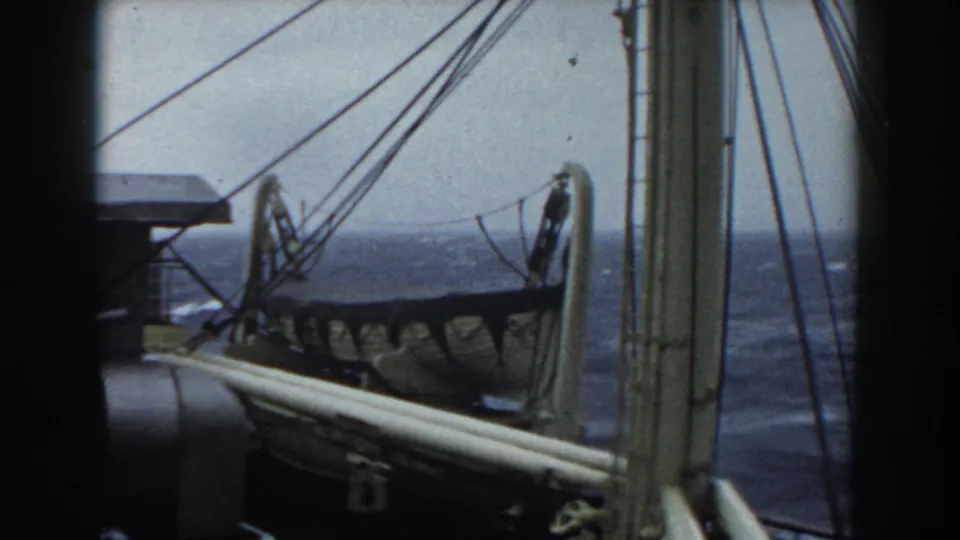 8mm frame from aboard a boat showing mast rigging, a deck rail, a side lifeboat, and dark Atlantic waves under gray sky.