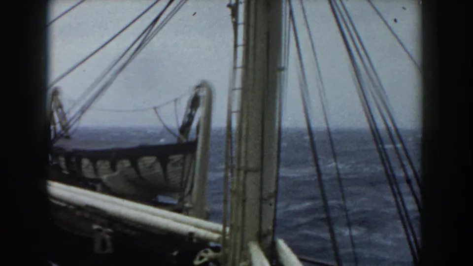 The camera looks past vertical mast rigging and a side lifeboat toward choppy open water with low gray sky.