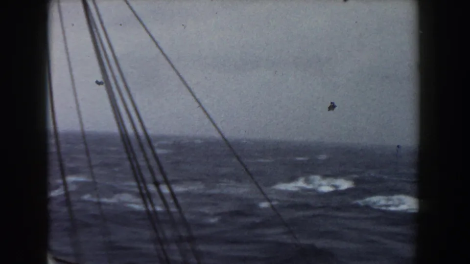 Boat rigging crosses the frame as the camera faces open Atlantic water with rolling swells, whitecaps, and a hazy gray horizon.