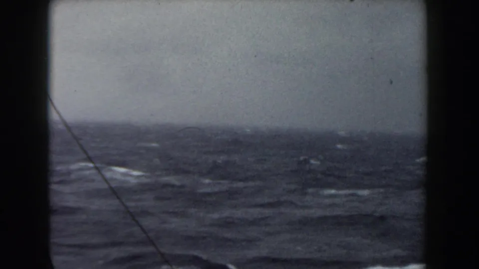 Stormy Atlantic swells under a gray sky, seen through the ship window with a diagonal rigging line in front.