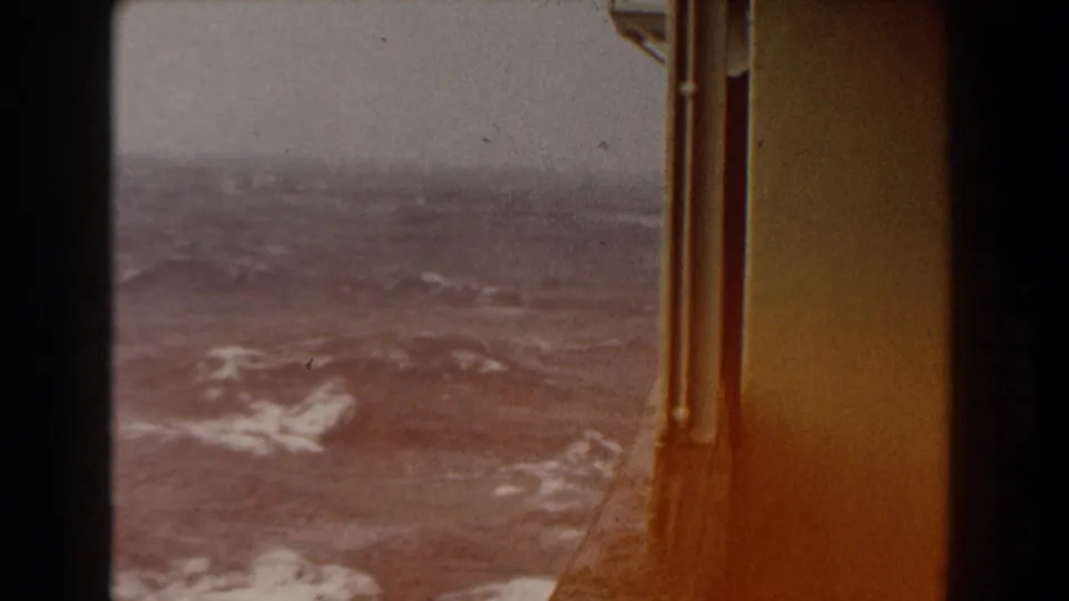 Choppy Atlantic water and white foam viewed past an amber-toned ship window or wall edge in the 1959 8mm footage.