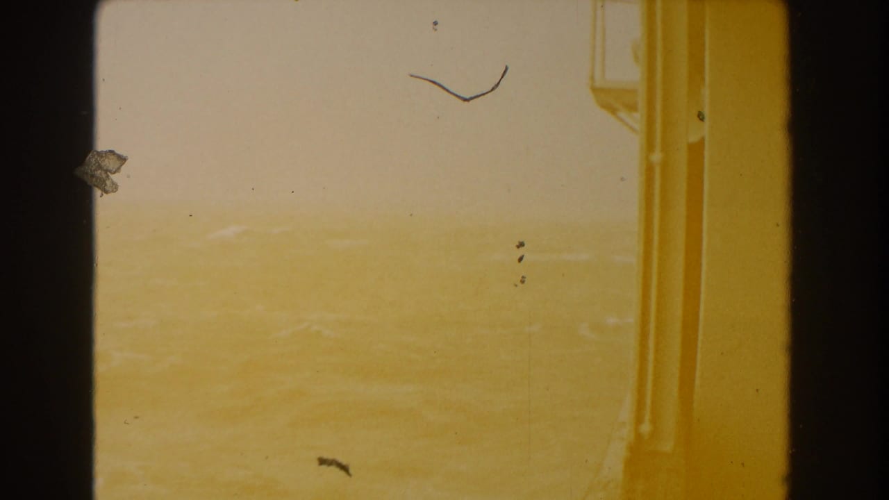 A hazy Atlantic Ocean horizon appears through a shipboard opening, with warm 8mm color cast, gate edges, and dust marks.