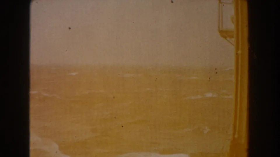 The sea horizon is faint through warm overexposure, bordered by dark ship structure and marked by small 8mm dust specks.