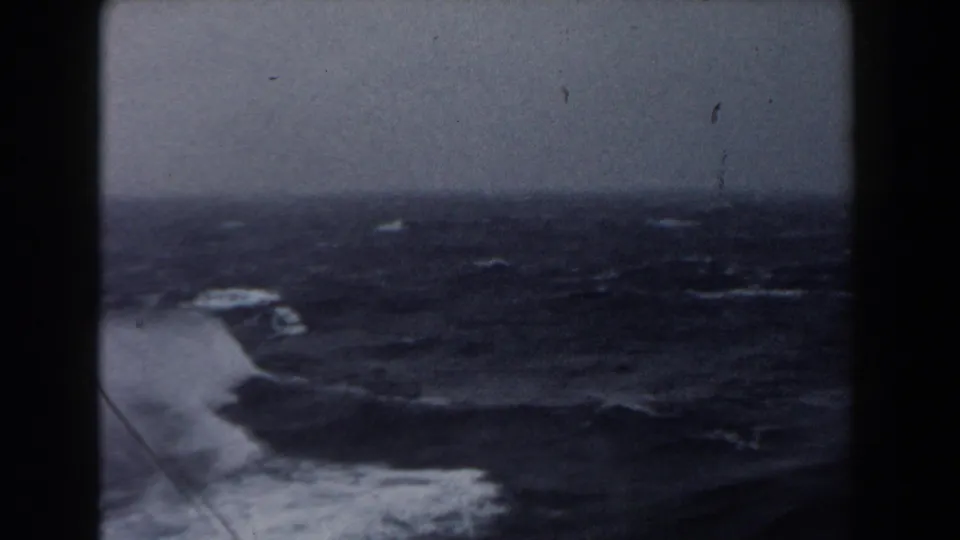 Rough dark ocean water churns beside the vessel, with white foam and spray visible near the lower left of the shipboard view.