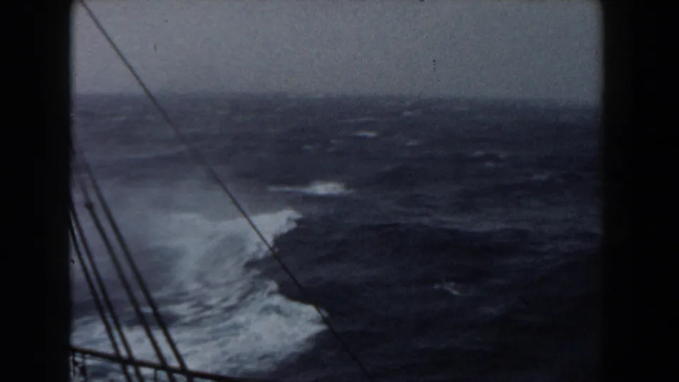 A wave breaks into white spray beside the ship, with dark water, a hazy horizon, and several rigging or cable lines crossing the frame.