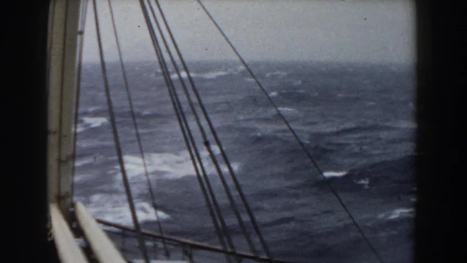 A mast and several rigging lines cut across a rough ocean view, with dark blue waves and gray sky visible through the film frame.