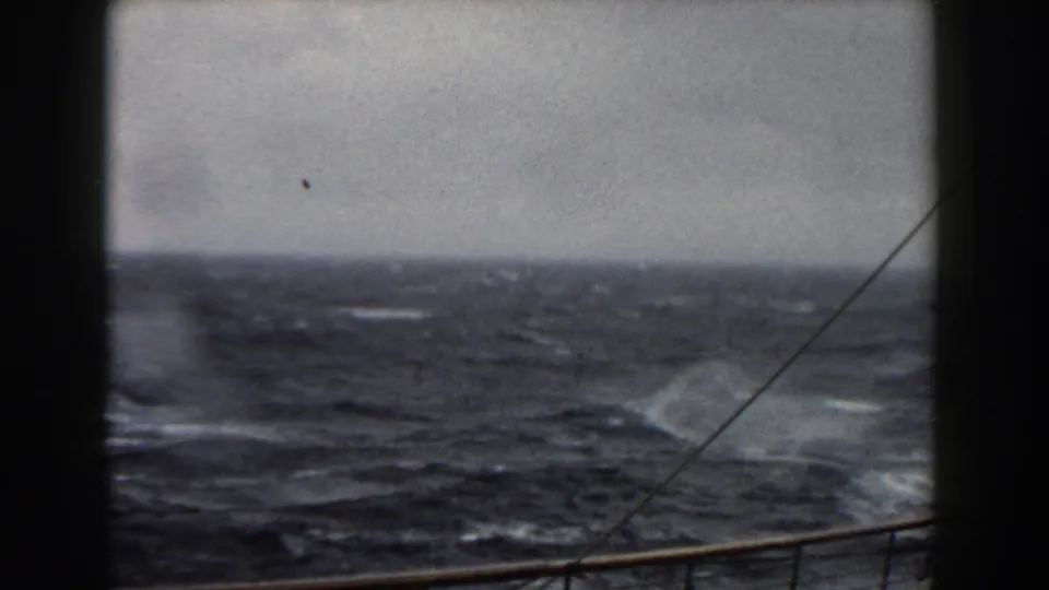 The camera looks over a low rail toward choppy open water, with whitecaps, haze, and a diagonal rigging line in the foreground.