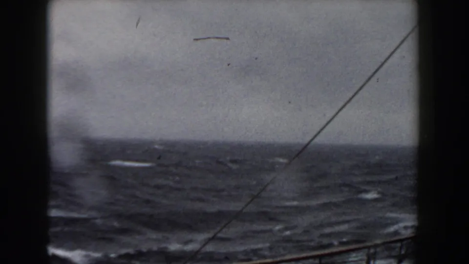 A rough gray-blue sea fills the frame, with whitecaps, a slanted rigging line, and the vessel rail visible near the bottom edge.