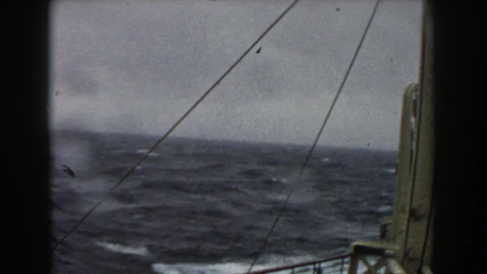 A handheld 8mm view from the ship deck shows rough ocean, whitecaps, rigging lines, and a vertical deck fitting at the right.