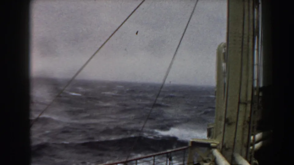 Choppy open water with whitecaps appears beyond ship railings, rigging lines, and a vertical vessel structure in the 1959 home-movie frame.