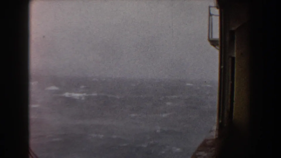 Distant whitecaps and a hazy ocean horizon are visible beside the dark vertical wall or opening of the ship in faded 8mm color.