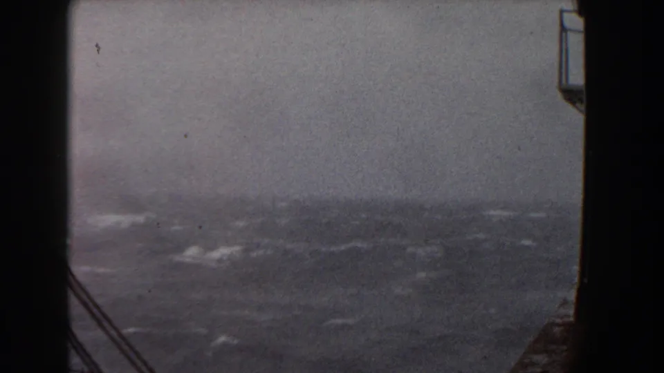 1959 8mm shipboard view of rough blue-gray ocean water under a cloudy sky, framed by dark vessel structure and rigging.