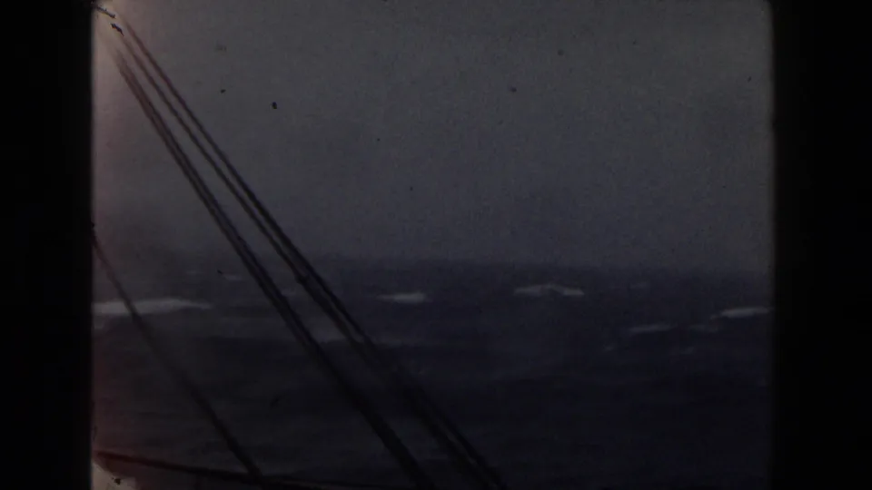 Grainy 1959 8mm frame showing diagonal rigging over dark blue ocean swells and scattered whitecaps in cloudy weather.