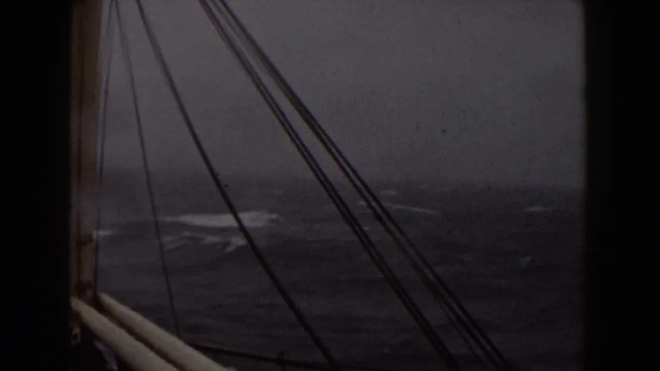 Late-1950s shipboard view of rough ocean water, with mast-like structure and rigging silhouetted against a muted cloudy sea horizon.