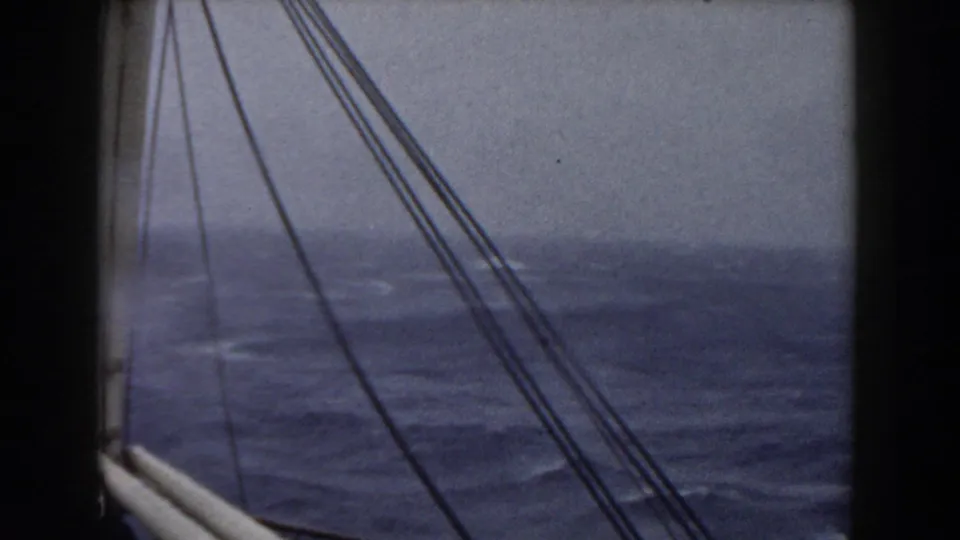 View from a sailing vessel across rigging toward gray-blue rough sea and a low, hazy horizon.