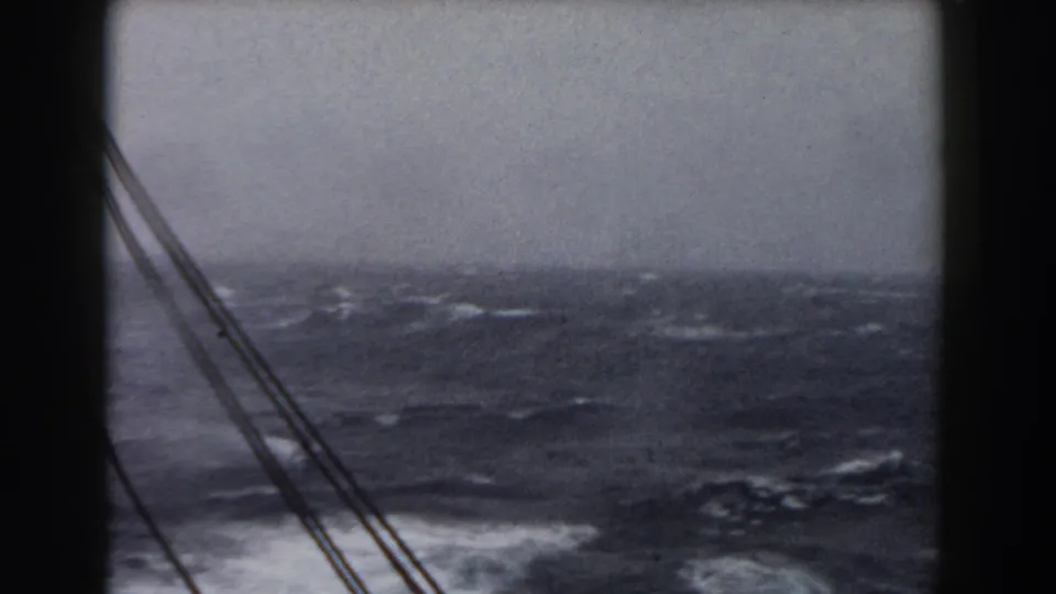 1959 8mm shipboard view of rough ocean water, whitecaps, overcast sky, and dark rigging lines in the foreground.