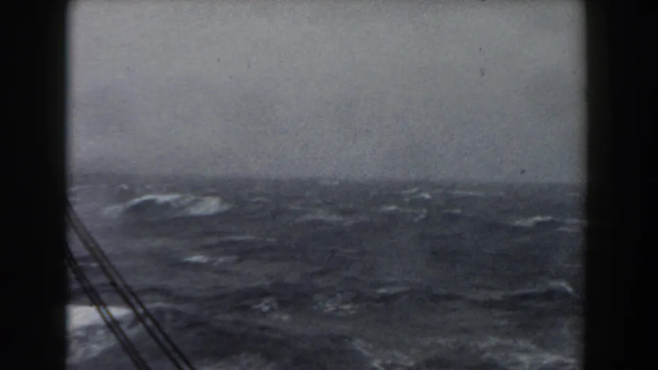 Shipboard 8mm view across rough Atlantic water, with whitecaps, a low gray horizon, rigging lines, and dark deck framing.