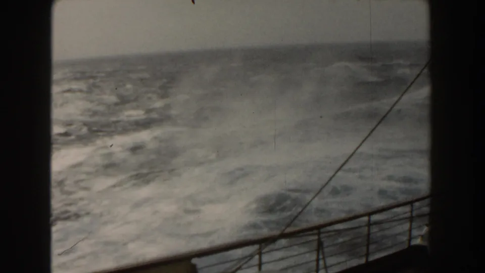 Rough Atlantic water churns beside the ship, with a foreground railing, diagonal rigging or cable, and softened 8mm color.